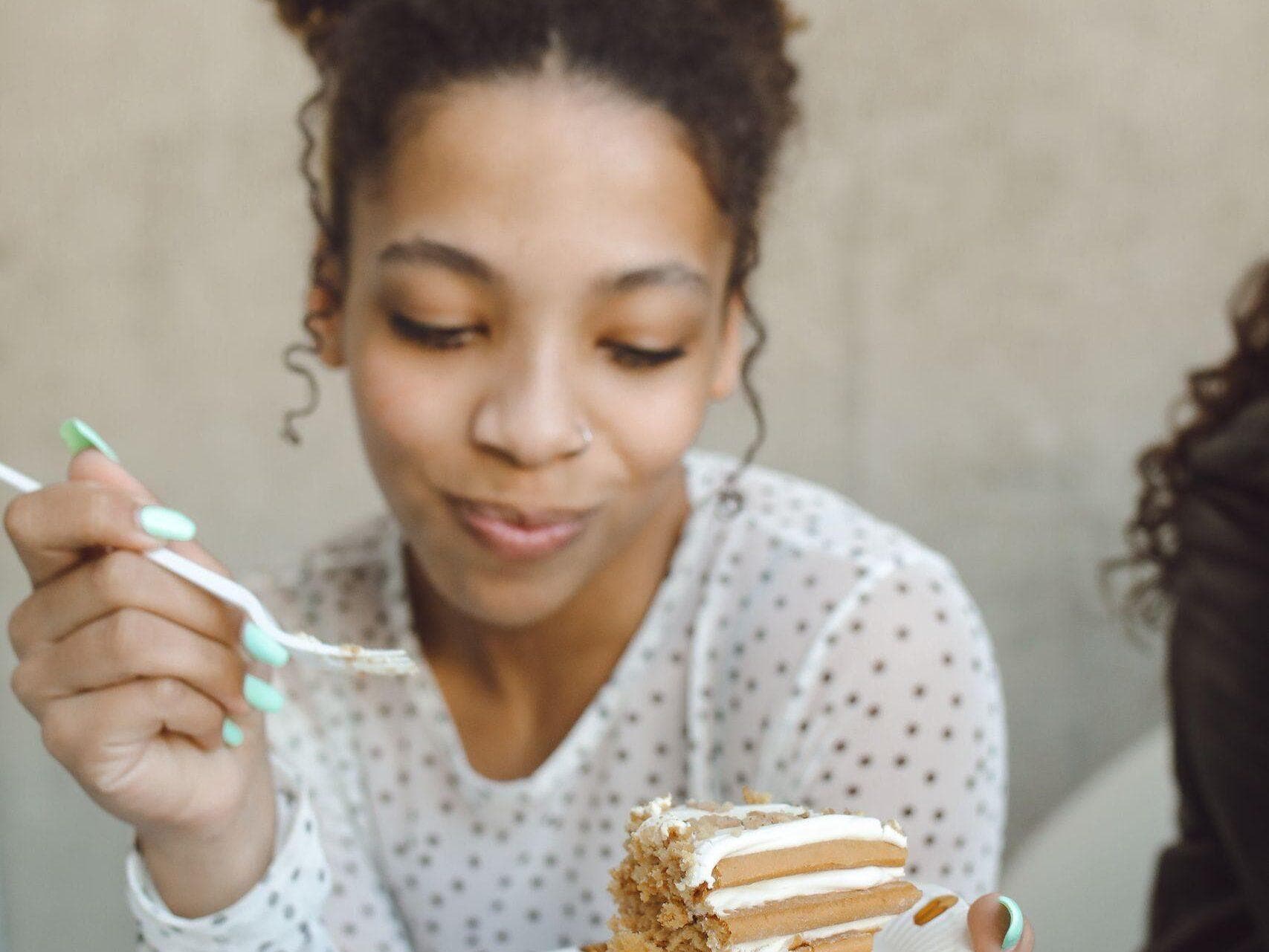 A cheerful young woman enjoys a slice of cake indoors, savoring a delightful moment.