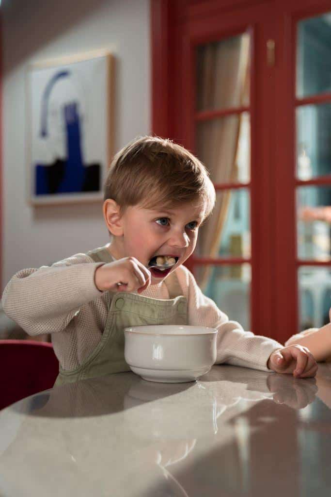 A young boy eating breakfast cereal with enthusiasm at home, seated at a kitchen table indoors.