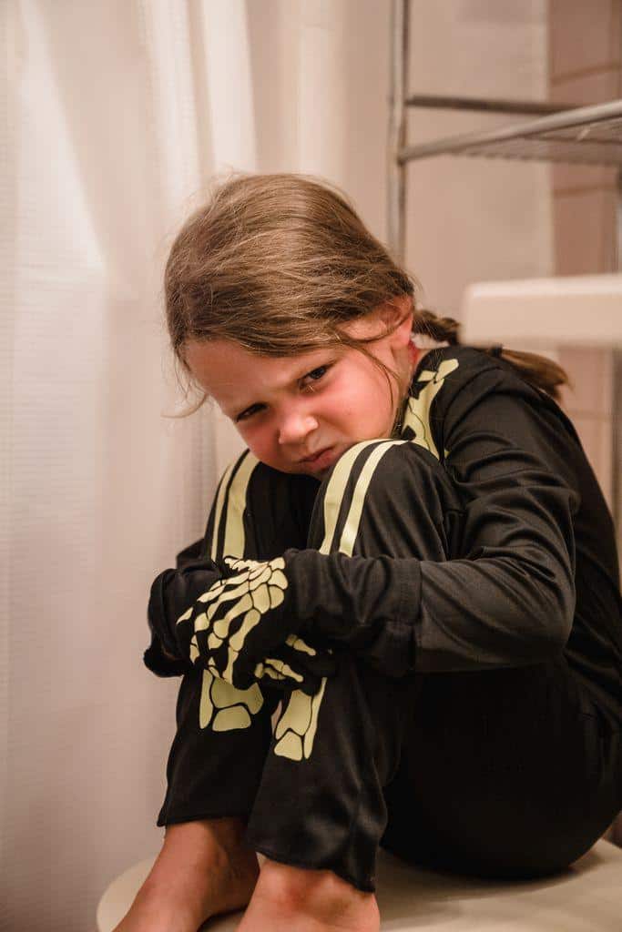 A young girl in a skeleton costume sitting indoors looking at the camera.