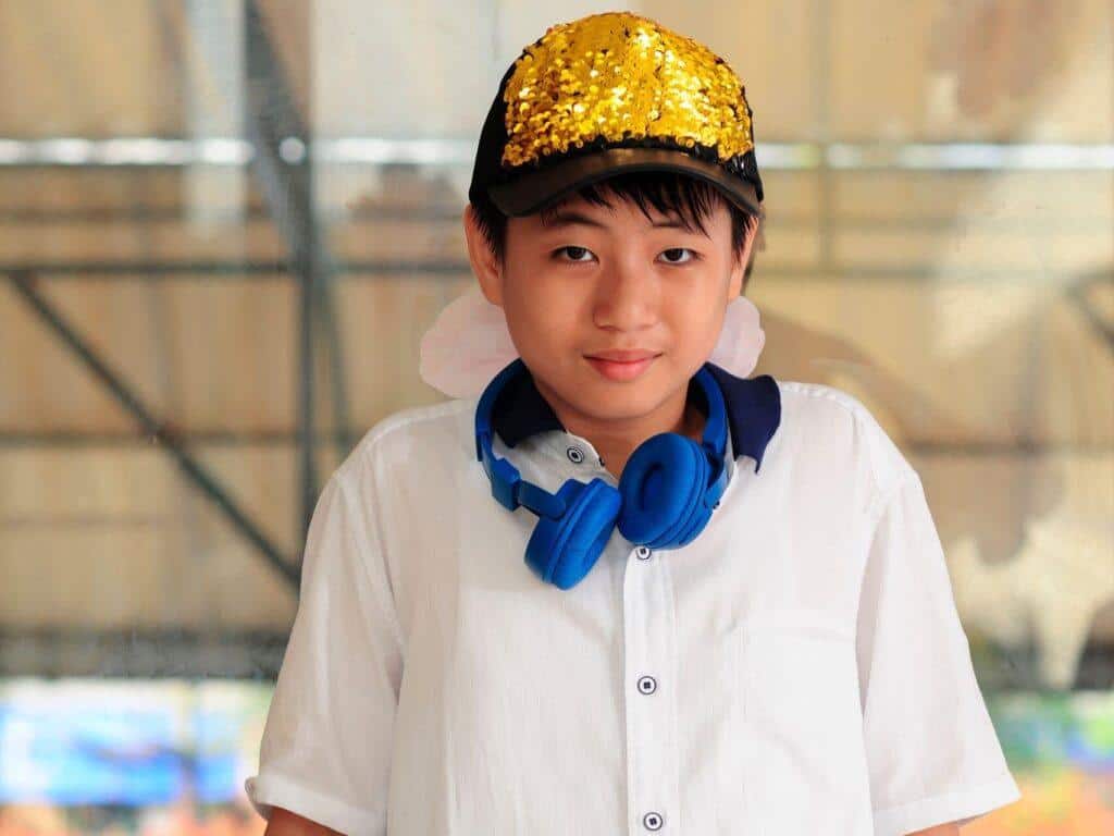 Portrait of a smiling Asian boy in a white shirt with colorful headphones and a sequin cap.